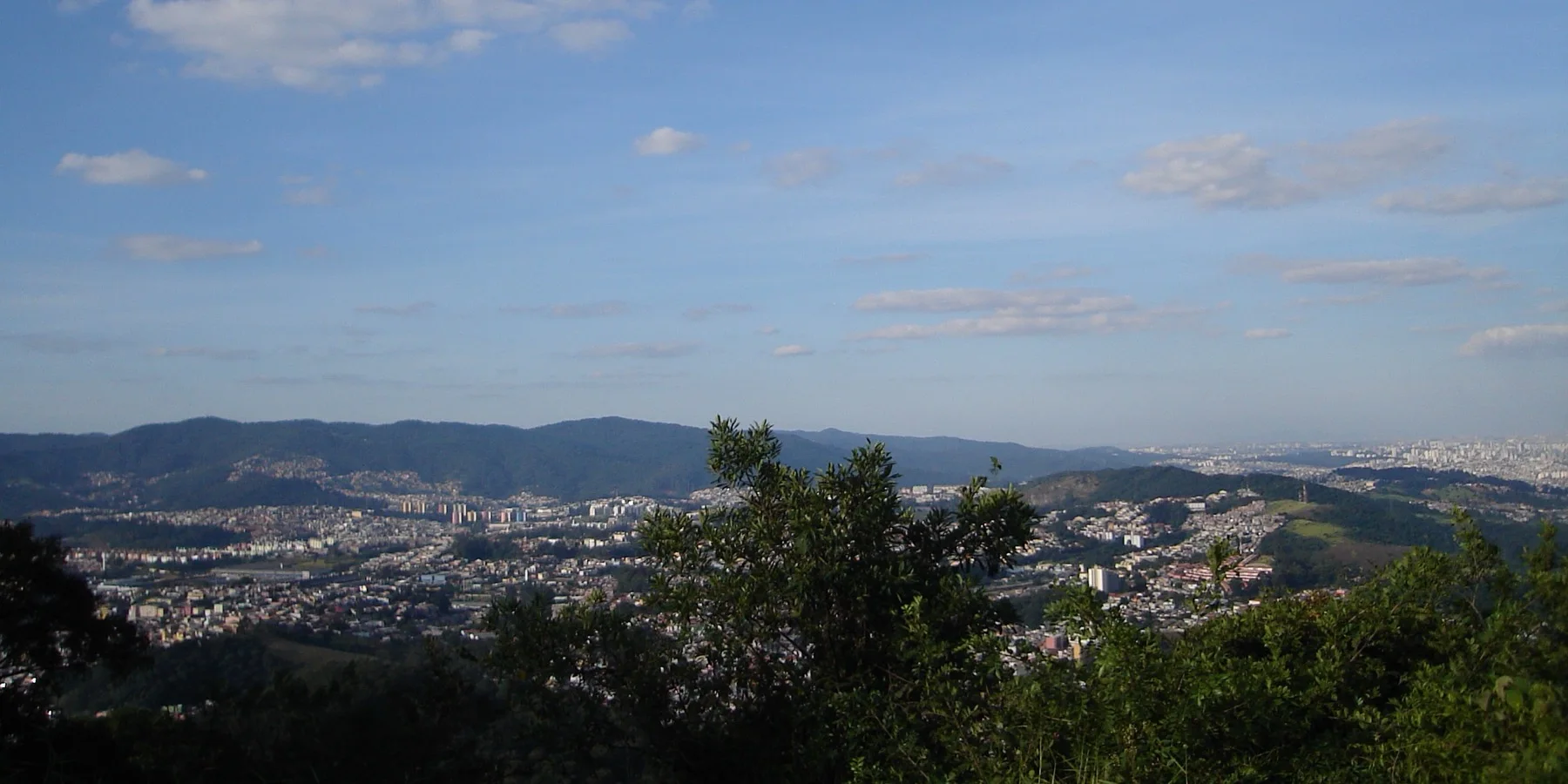 A cidade de São Paulo vista do alto de uma encosta de mata, com a vegetação em primeiro plano e a mancha urbana se estendendo entre morros até o horizonte