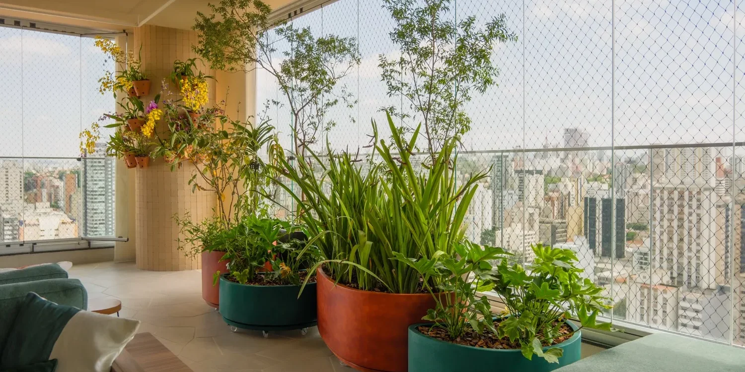 Varanda de apartamento em São Paulo com floreiras de terracota e verde, orquídeas na parede e o skyline da cidade atrás da tela de proteção